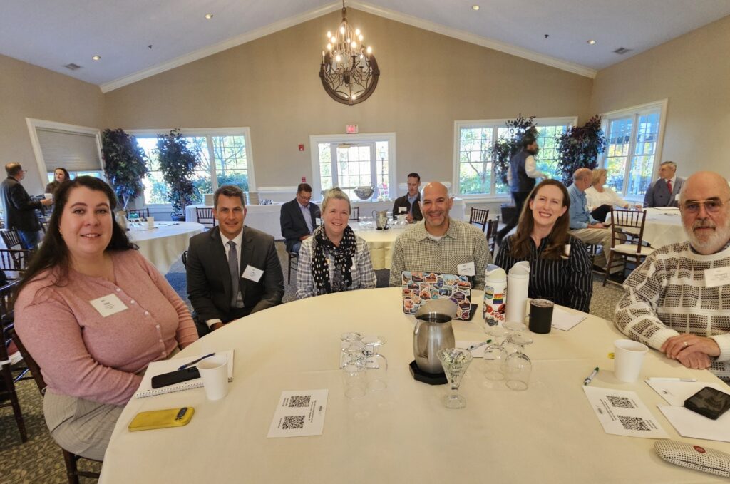 A number of people sit around a table in what appears to be a ballroom.