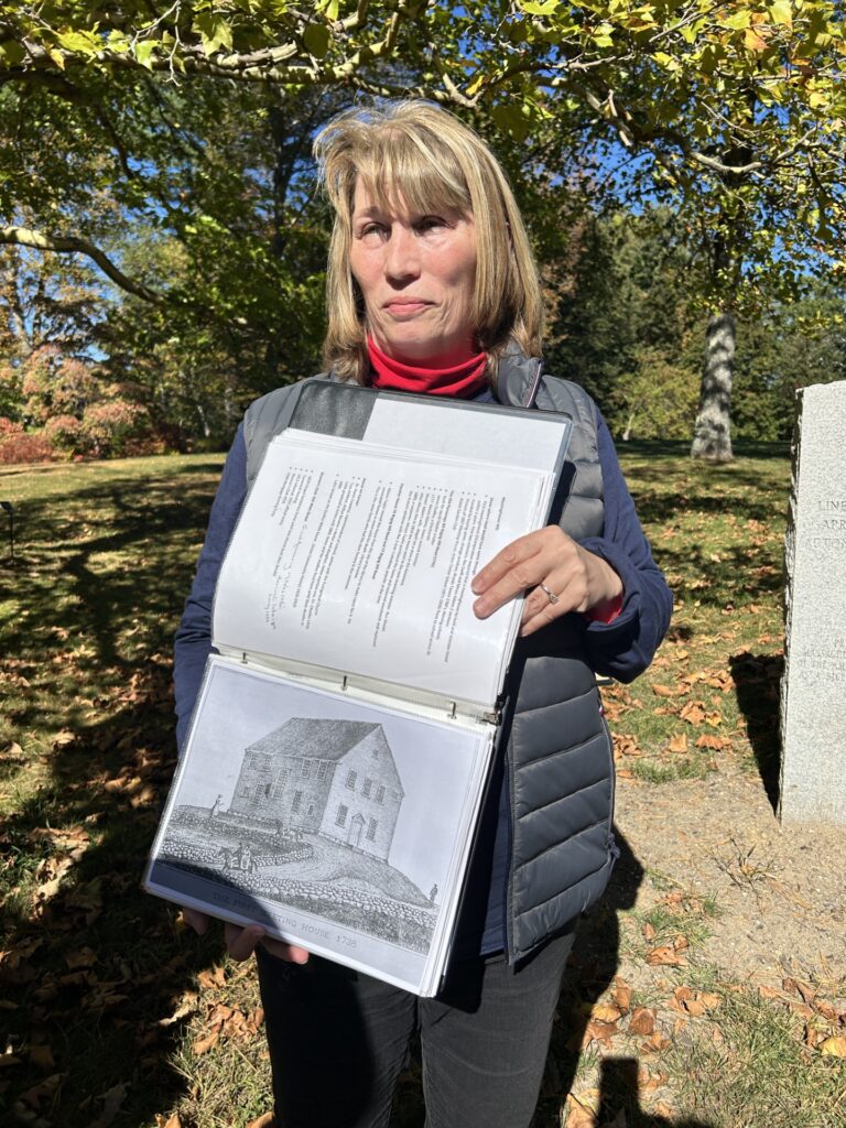 A blonde woman holds a notebook that shows a drawing of the original Acton Meeting House.