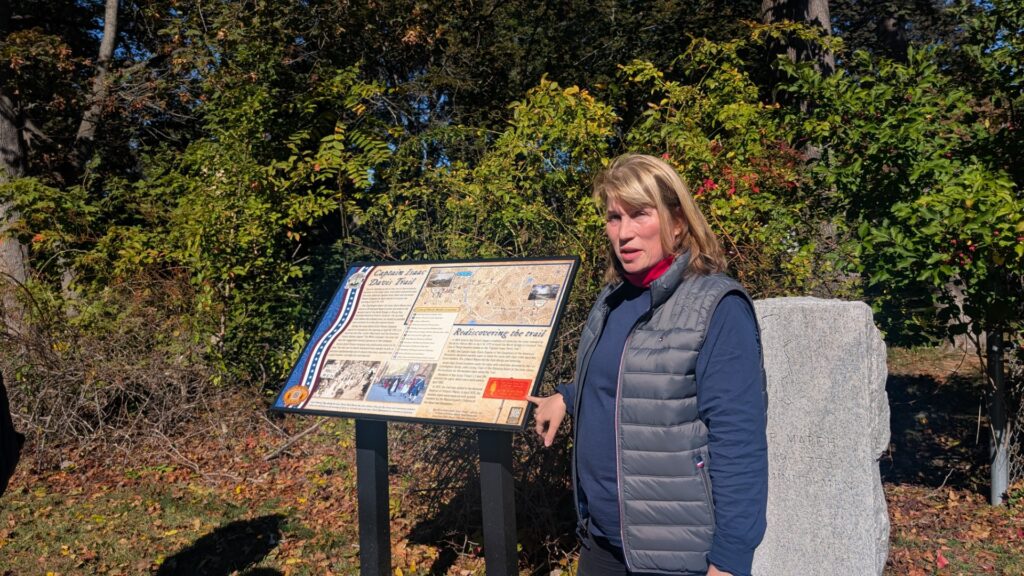 A blonde woman stands next to an information panel that marks the Isaac Davis trail. Behind her is a stone pillar that was installed in 1975.