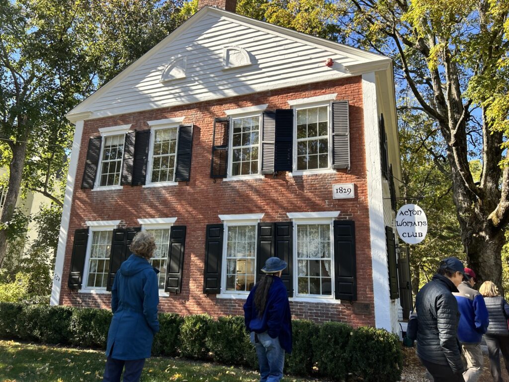 People walk into a brik and wood building that has the date "1829" on the wall and an Acton Woman's club sign on the side.