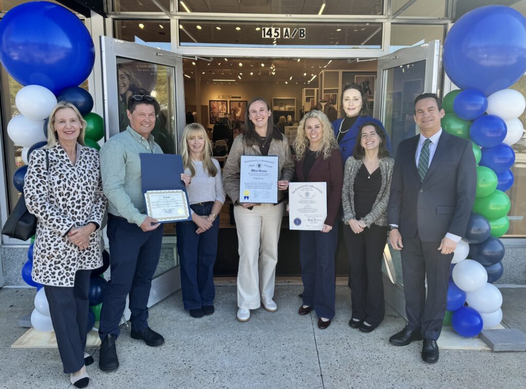 A group of people stand in front of an open doorway. There are blue, whit, and green balloons on both sides of the door, and some people are holding citations.
