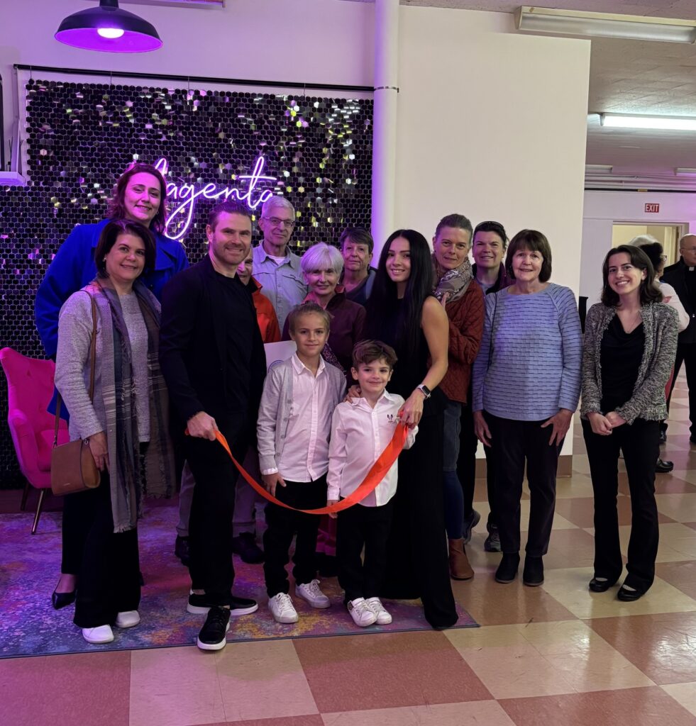 A group of people, including some children, stand in front of a sign that says Magenta/ Two people are holding a red ribbon and the entire picture is lit with a purplish (magenta) lght.