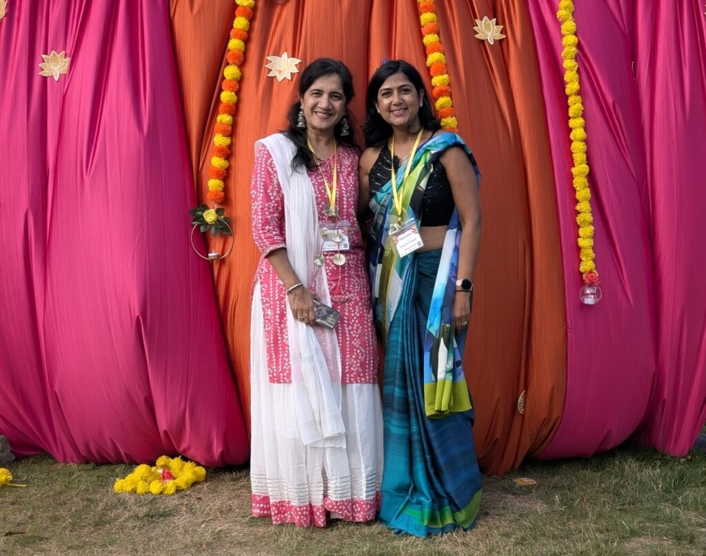 Two women wearing Indian clothing stand in front of a brightly colored curtain.