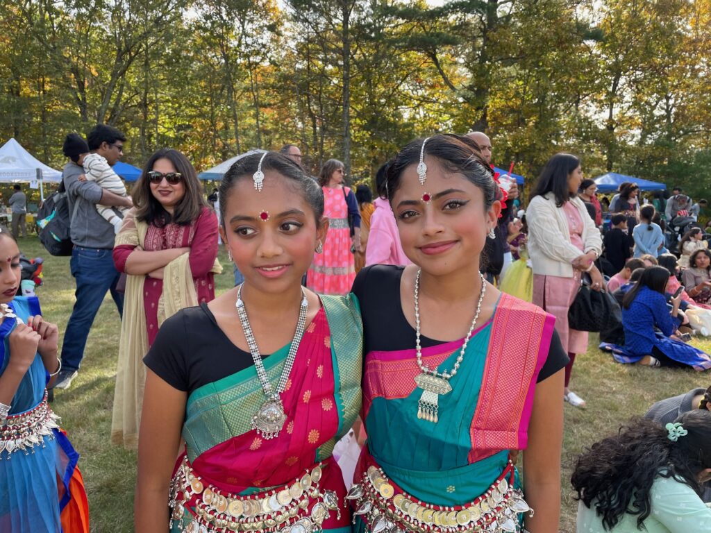 Two young woen wearing Odissi dance costumes and lots of silver jewelry stand next to each other. In the background, people are walking around or sitting on the grass to have lunch.