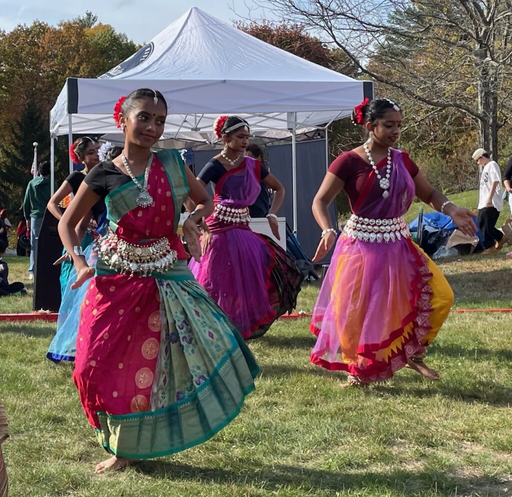 Several barefoot young women dance wearing brightly colored outfits and jingly silver belts.