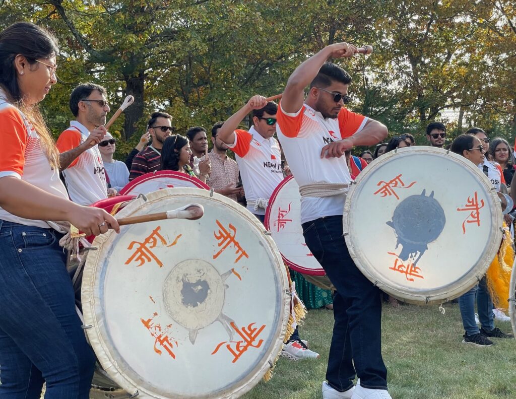 A group of drummers in orange and white shirts beat on drums. One of the drummers has his hands in the air, looking very dramatic.