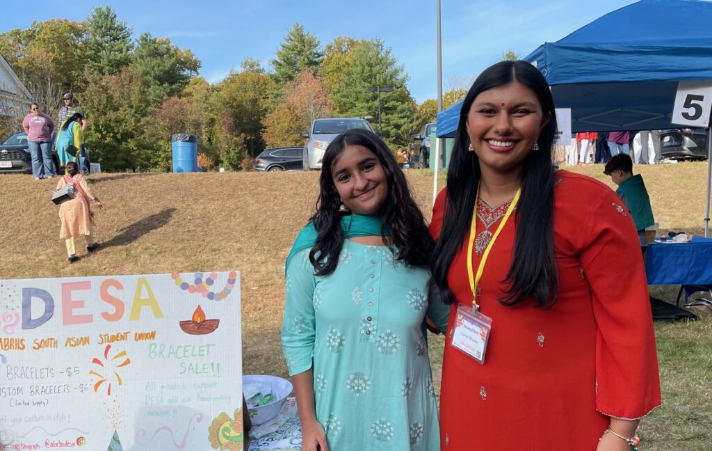 Two young women wearing salwaar kameez stand next to a DESA poster.