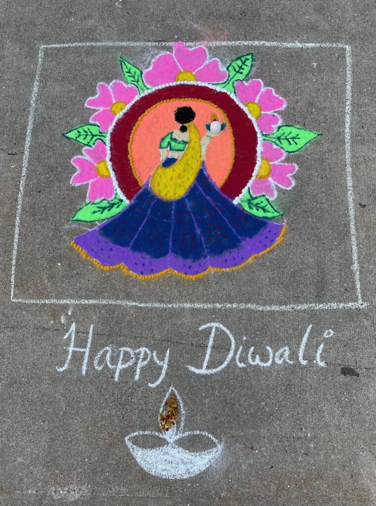 A sidewalk drawing of a woman in a purple skirt carrying a lamp. She is surrounded by a circle of flowers and walking into an orange circle. Underneath are the words "Happy Diwali" and a picture of a little oil lamp.