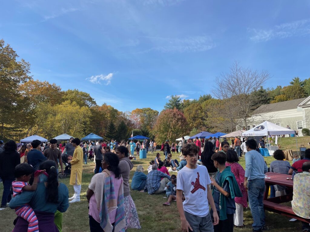 People in both western and south Asian clothing walk around a sunny field and sit on the grass. There are tents in the background selling food and other items.