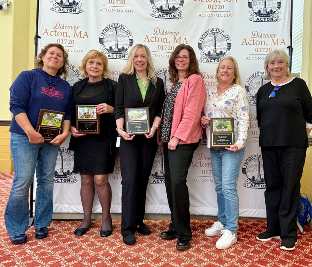 A group of women stand in front of a sheet with the Town of Acton logo (and discover Acton, MA design). Many of the women are holding the plaques that they were just awarded by the Acton Garden Club.