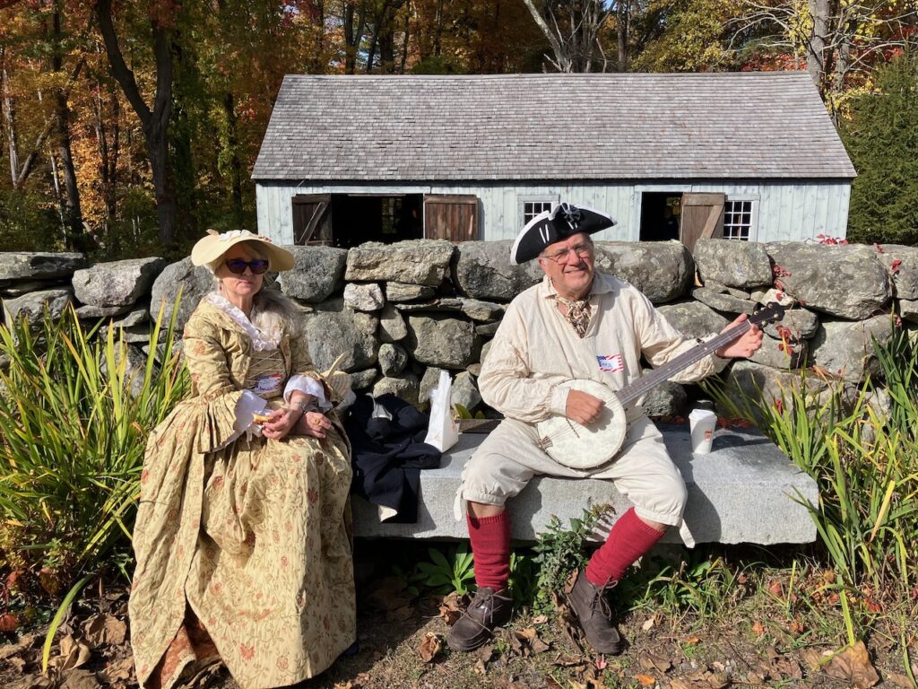 Two people, a man and a woman, sit on a stone bench in front of an old barn. The man is anachronistically playing a bango.