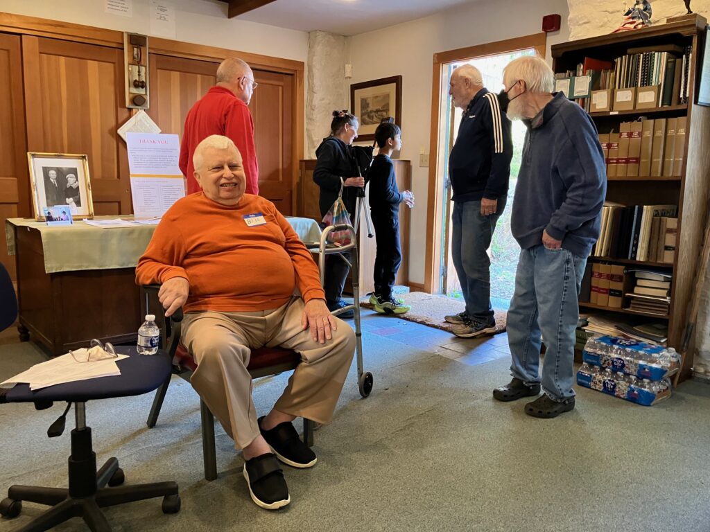 A man in an orange shirt sits at a chair inside an old house. A numbner of other people are looking around the house or out the door.