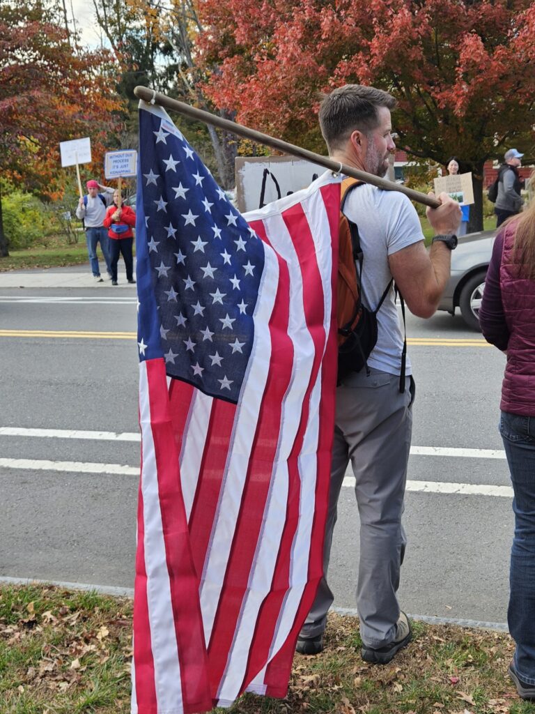 A man carries a large American flag over his shoulder on a wooden pole. He is standing by the street so drivers can see it as they go by. Across the street are a number of other attendees, many with signs.