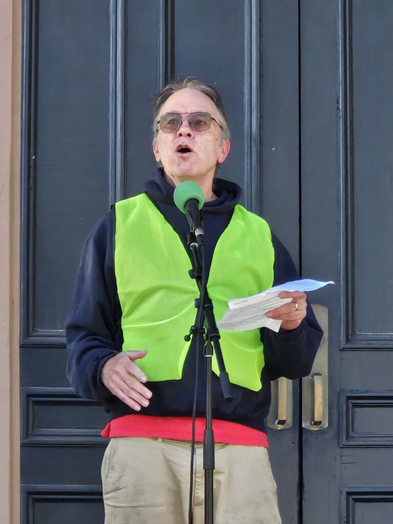 A man wearing a bright yellow marshal vest speaks into a microphone on the steps of Town Hall.