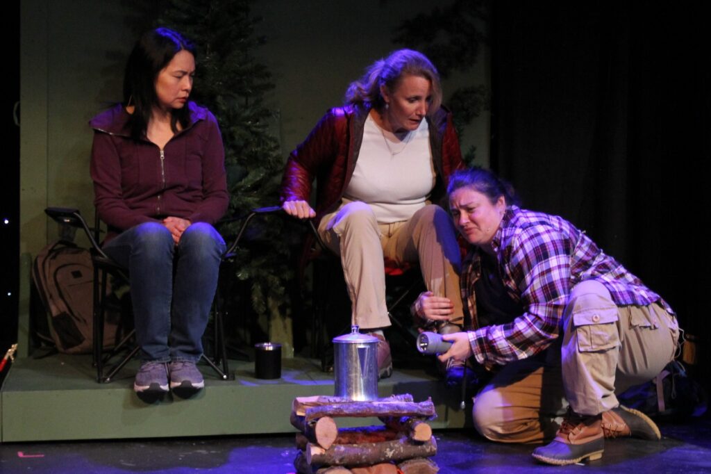 Two women sit in camping chairs in front of a wood fire. Another woman is on the floor hugging the leg of one of the seated actors.