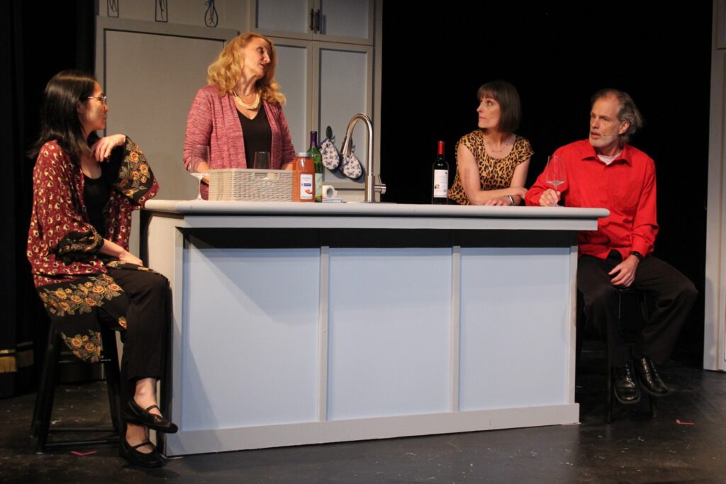 Three women and a man surround a kitchen counter onstage. Three of the actors arre sitting with glasses of wine; a fourth is standing in front of a sink.