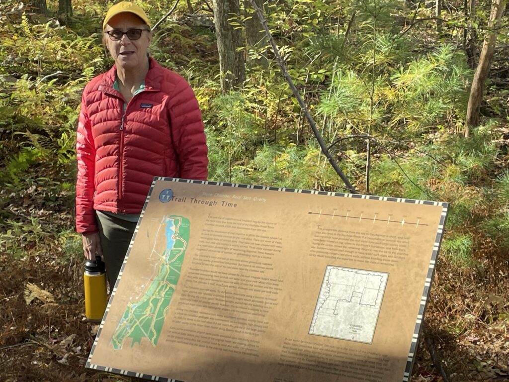 A woman with a red jacket and yellow hat stands next to a Trail Through Tiime trail marker.
