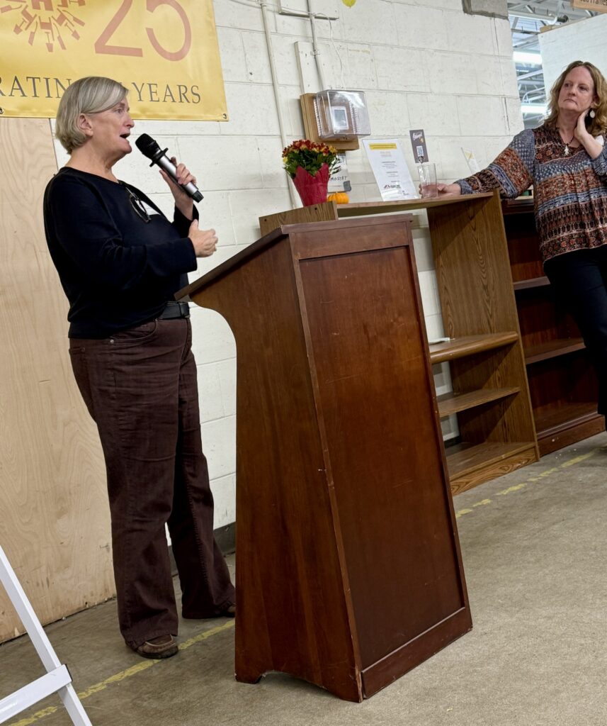 A woman with shortish grey hair speaks at a podium. Another woman listens in the background.