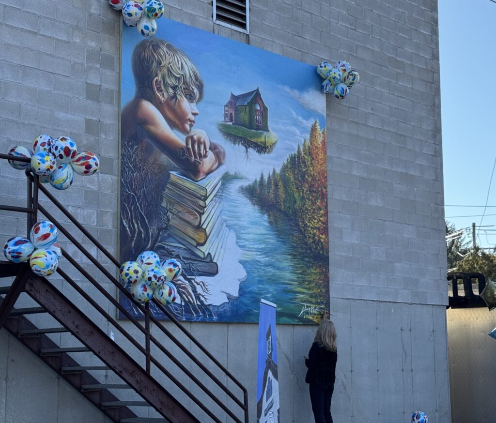 A woman signs the very bottom of the mural. There are bunches of multi-color balloons in the corners and on the stairs in front of the mural.