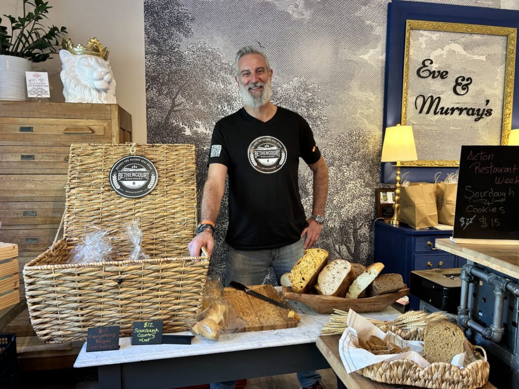 A tall bearded man stands behind a large basket that contains bread. There are sliced loaves available for tasting on the table.