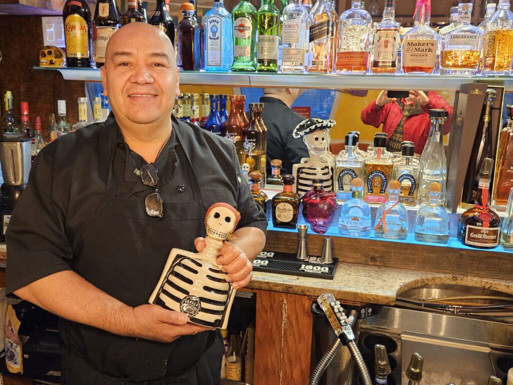 A bald, smiling man stands in front of a liquor shelf. He's holding a skeleton shaped bottle (labeled Skelly).