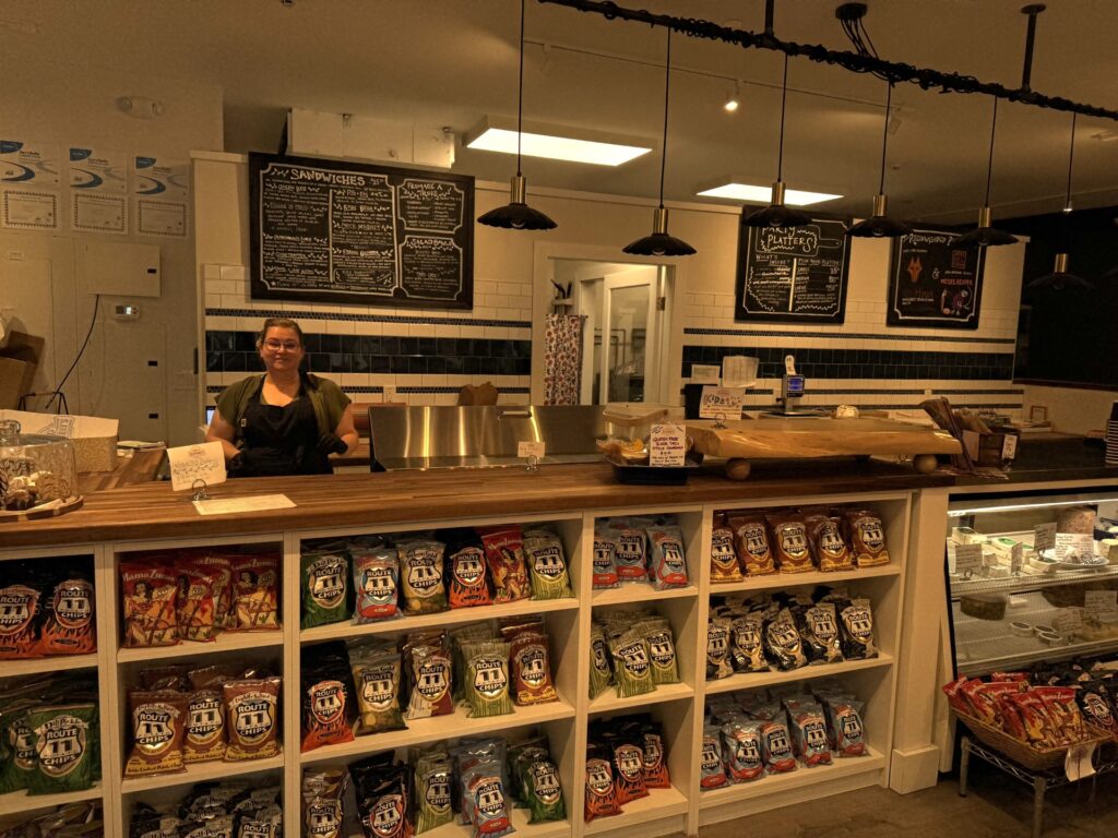 A woman stands behind a service counter. On the background a blackboard lists all of the sandiches. On the right, you can see the cheese cooler, chock full of tasty delights.