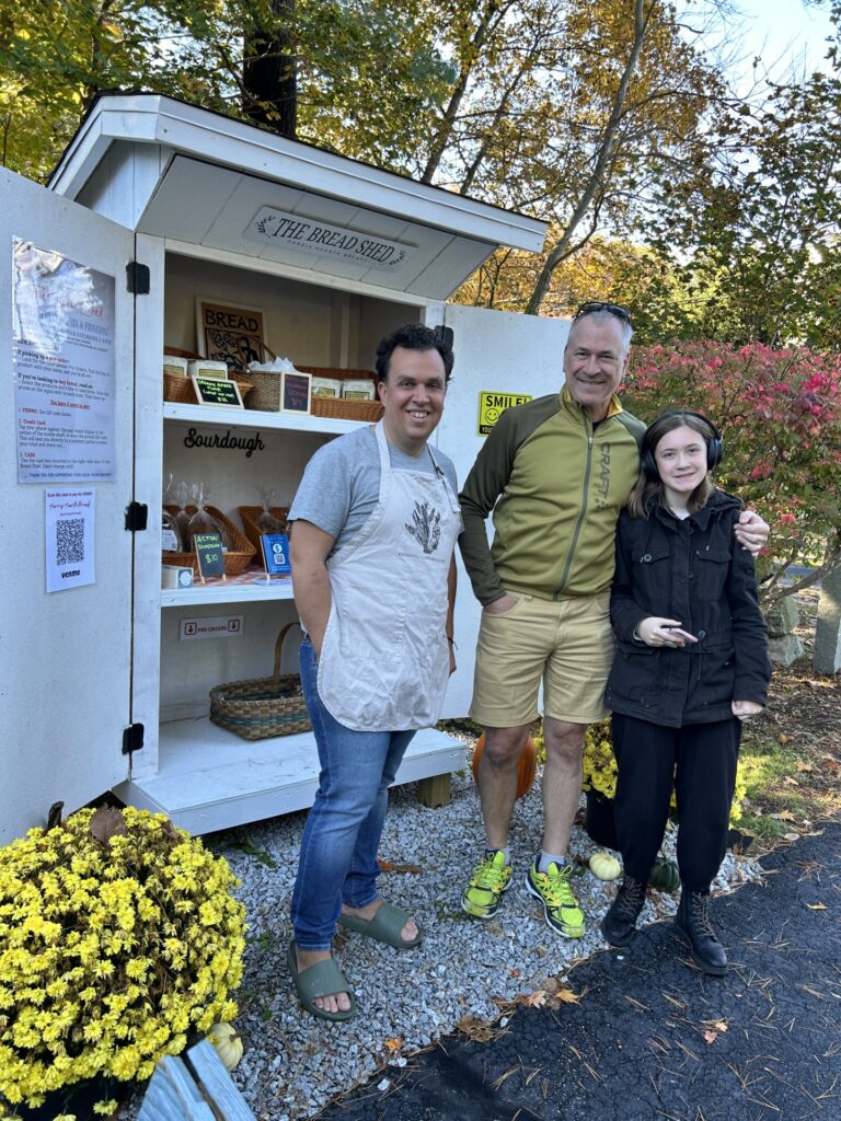 Three people stand in front of a small building (a shed). The man on the right, the owner, wears an apron. The shed contains breads and other pastries for sale or for pickup by customers who pre-ordered.