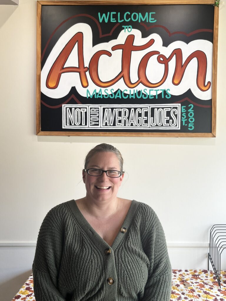 A smiling woman stands under a Not Your Average Joe's sign that reads "Welcom to Acton, Massachusetts."