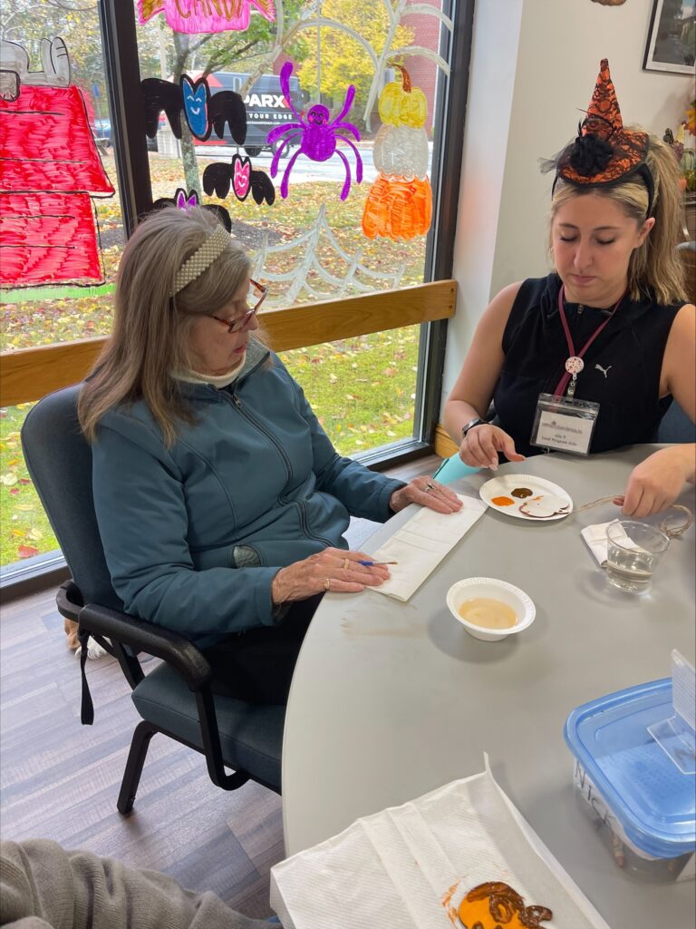 A young woman wearing a pointy orange hat (with a spider) helps an older woman set up a halloween craft (decorating cookies, perhaps).