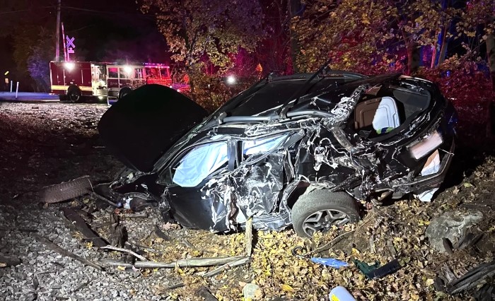 side view, at night, of a car – smashed and crushed, suronded by debris. A red fire engine is in the background.