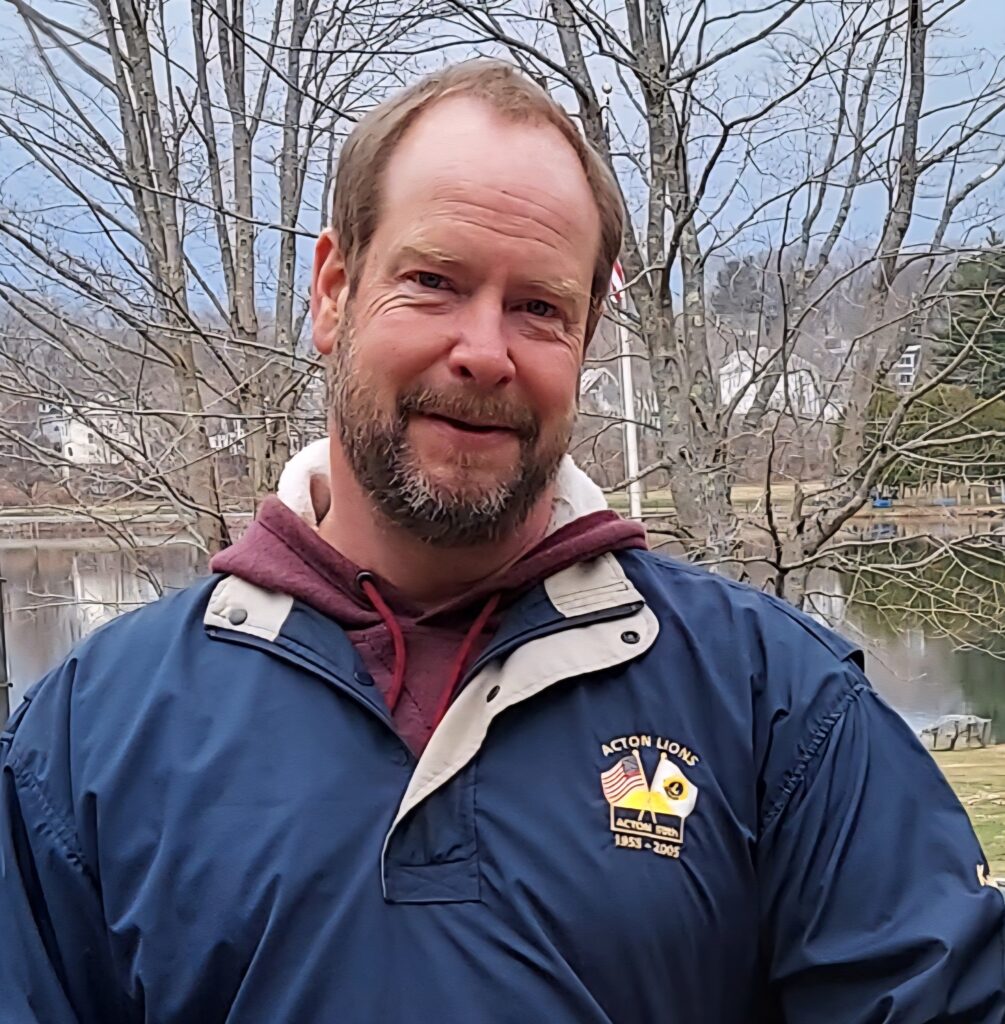 A beared man in a blue parka stands before bare trees.