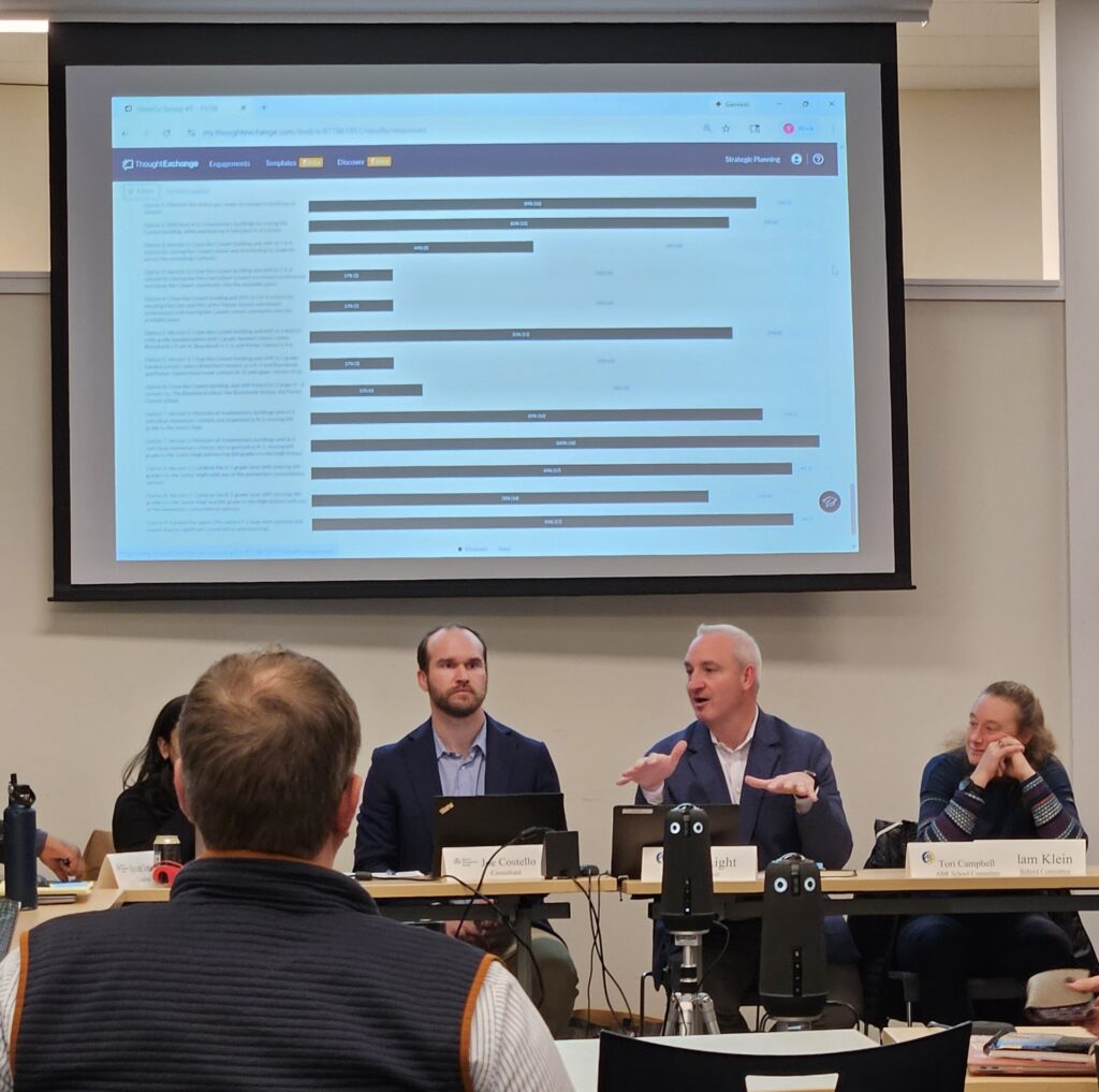 Several people site at a table (and the head of an audience member blocks some of them). A silver haired man is sitting at the table and speaking.