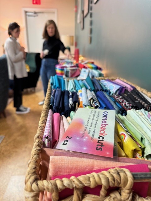 Baskets of fbrightly colored abric (fat quarters) line a wall. In the background, one woman is describing something to another woman.