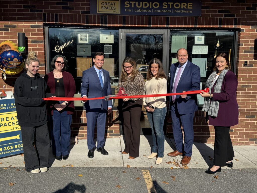 A group of people holding a red ribbon in front of a store. One woman is cutting the ribbon with Acton's oversized shears.