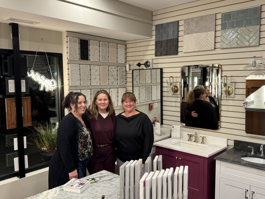 Three women pose in a room with tile samples on the back wall, and various sinks and cabinets in the background.