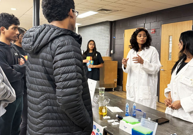 High school students talk to a couple of adults wearing lab coats. On the table are various tools of the trade, including test tubes and pipettes.