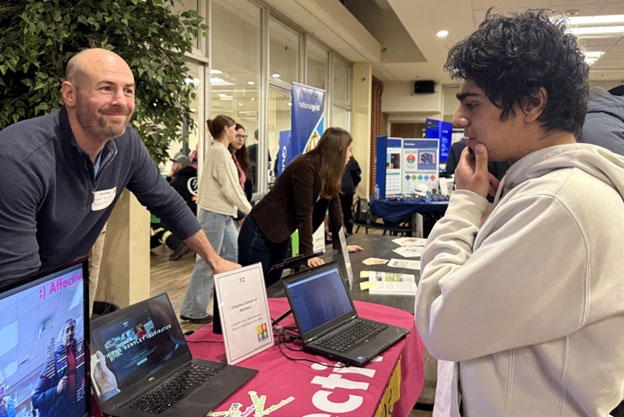 A student is looking at a computer screen. On the other side of the table, a salesman is leaning over waiting, perhaps, for questions.