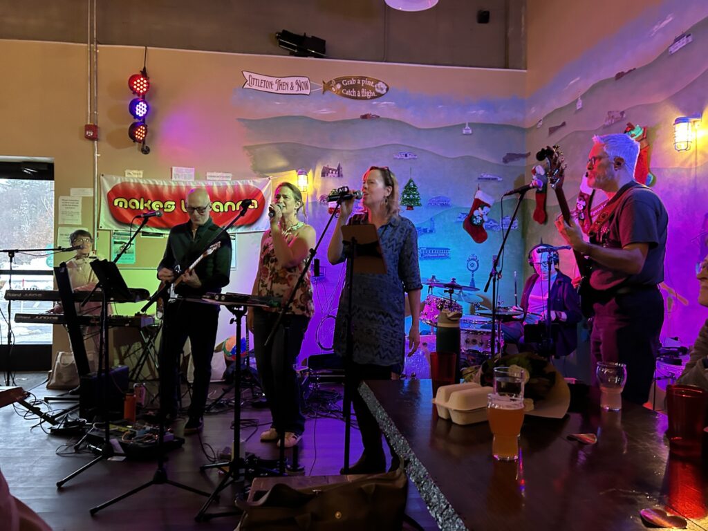 A band in a stage area wtih multi-colored lights and a "makes u wanna" sign on the back wall.