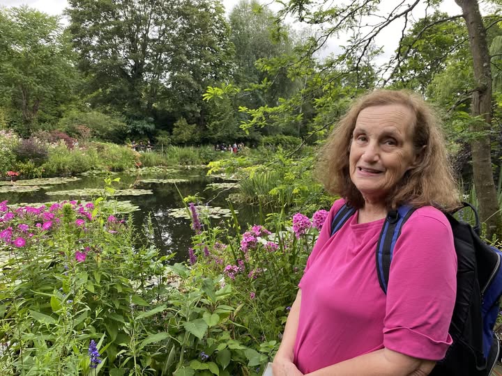 A smiling woman in a pink shirt stands in front of a pond. Right behind her are plants and flowers, many of which match her shirt.