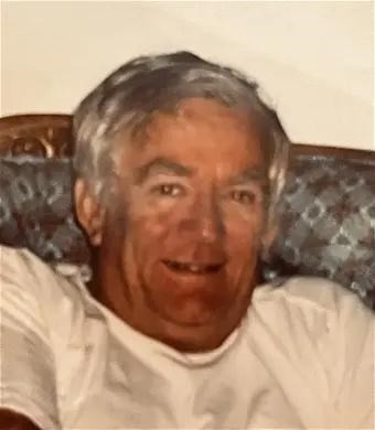 Aman in a white shirt leans against a couch.