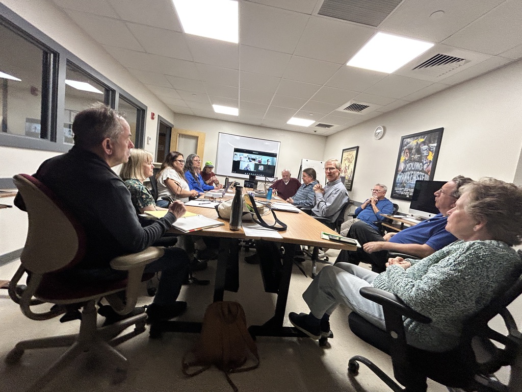 A group of people sitting around a table. In the front is a computer screen.