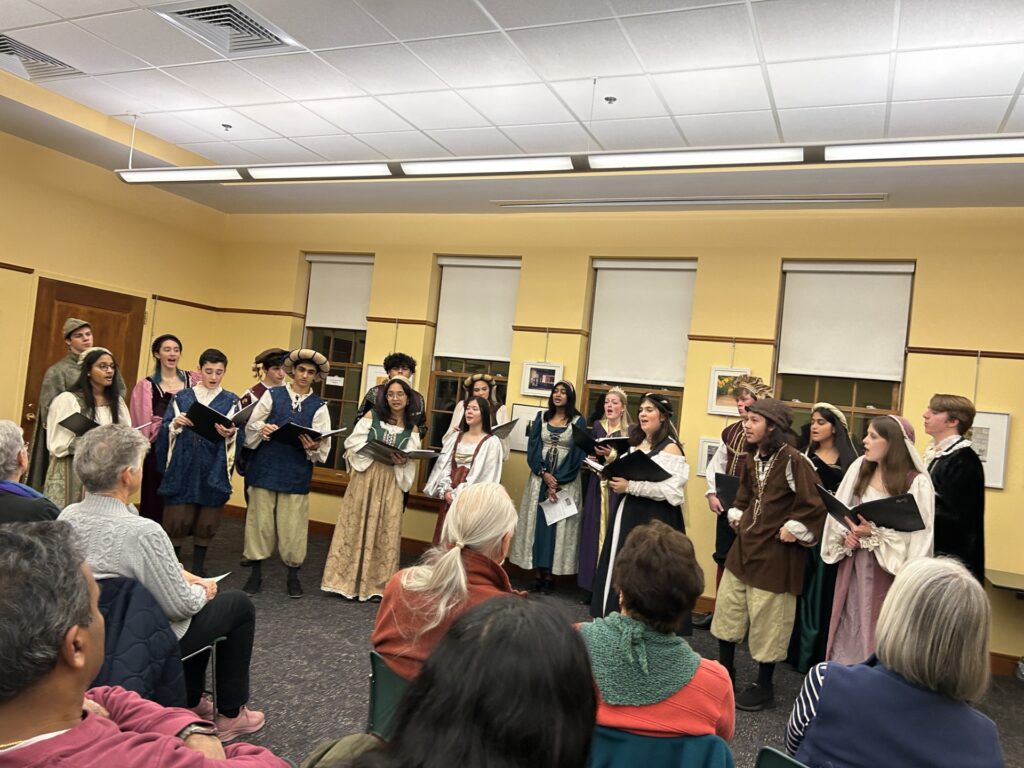 A group of high school singers, in medieval garb (including great hats!), sings in the Sargent Library meeting room.