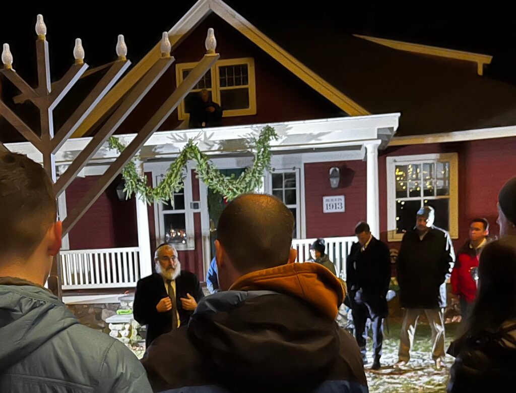 A bearded man stands in front of the Red House, and under a tall menorah, and speaks.