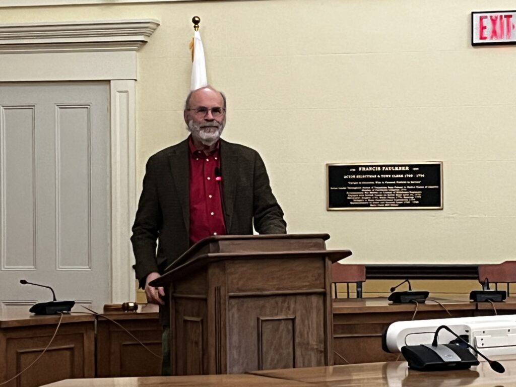 A bearded man wearing a red shirt and a jacket speaks at the podium in Town Hall.