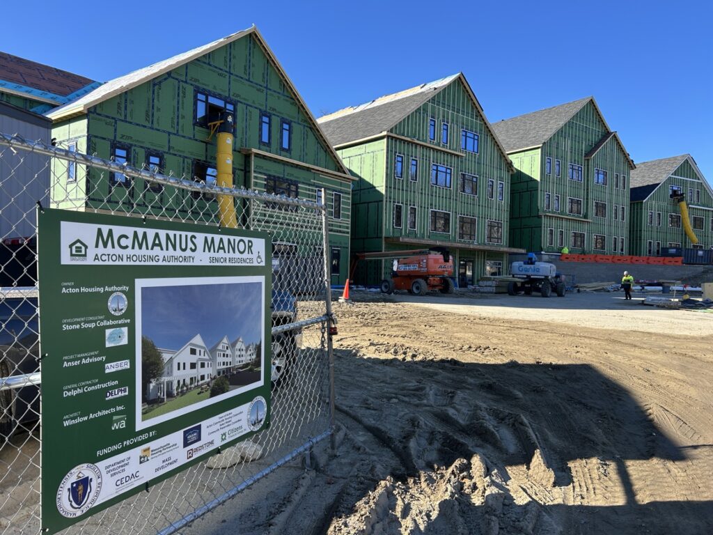 Four Tyvek-covered buildings stand in the sun. In the foreground, a gate with an architectural rendering of the buildings, labeled McManu Manor.