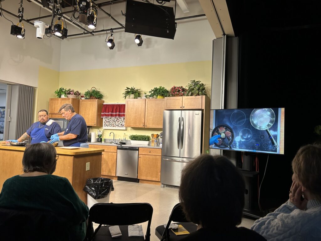 Two men work in a brightly lit show kitchen. On the right, a screen shows what's going on on the stove. There are audince members seated around the kitchen.