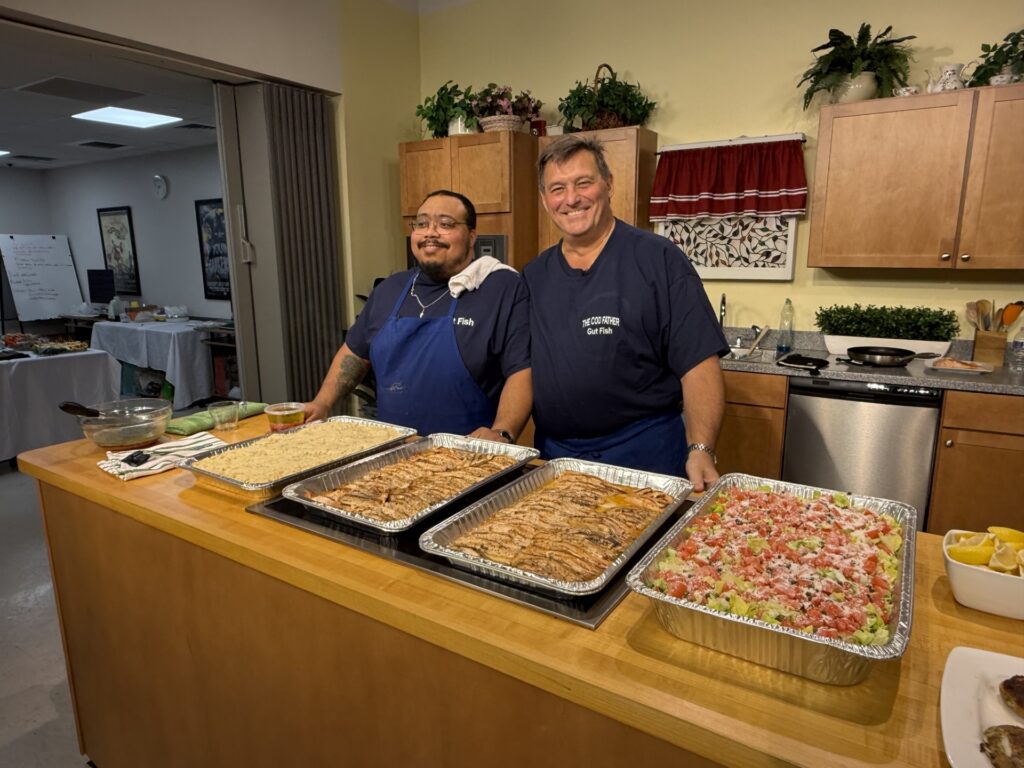 Two men stand in front of a counter full of delicious-looking food: rice, salmon, and salad.