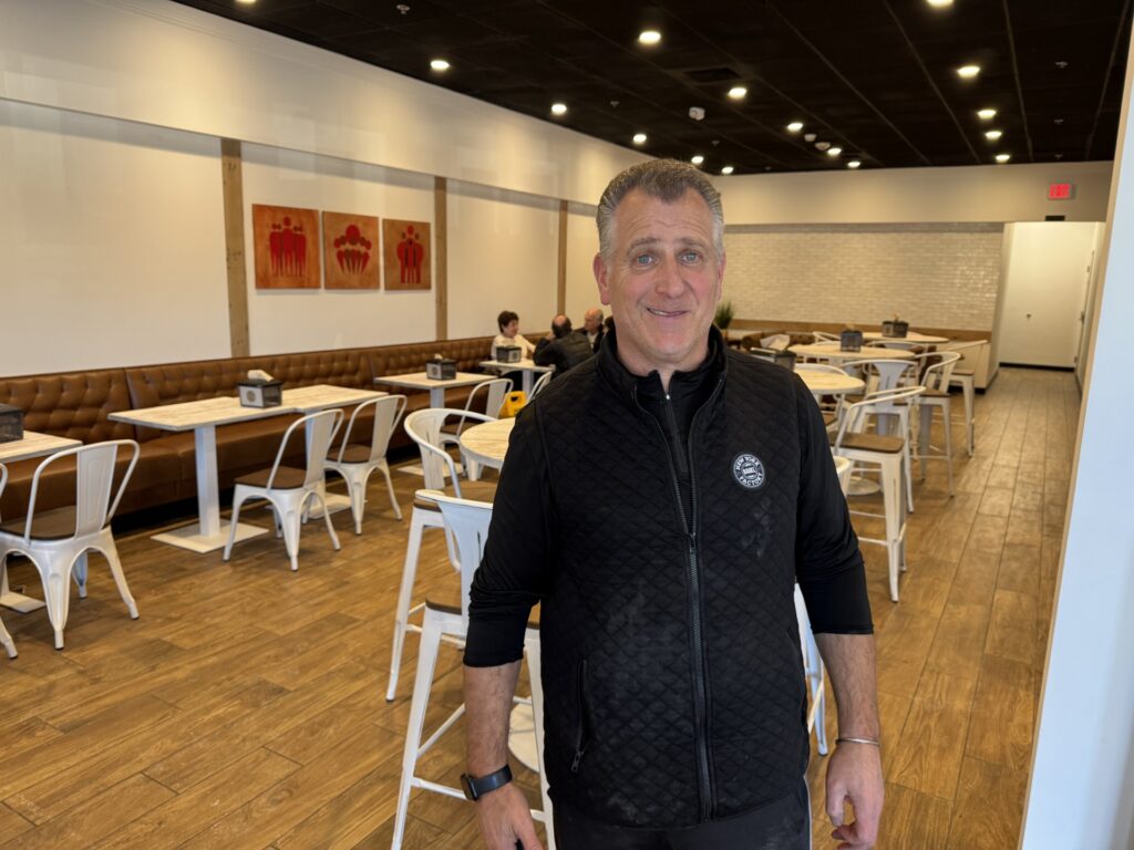 An older man in a black sweatshirt stands in front of an almost empty restaurant.
