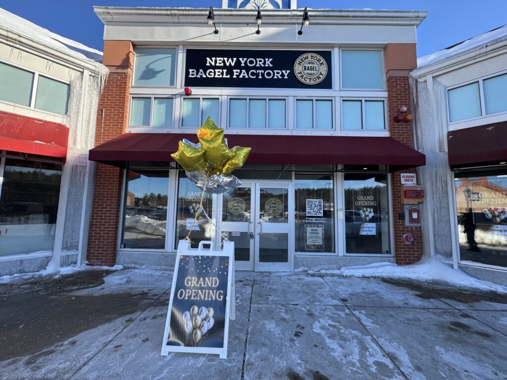 A corner building with a large New York Bagel Factory sign over the door. In front is a Grand opening sign with some balloons.