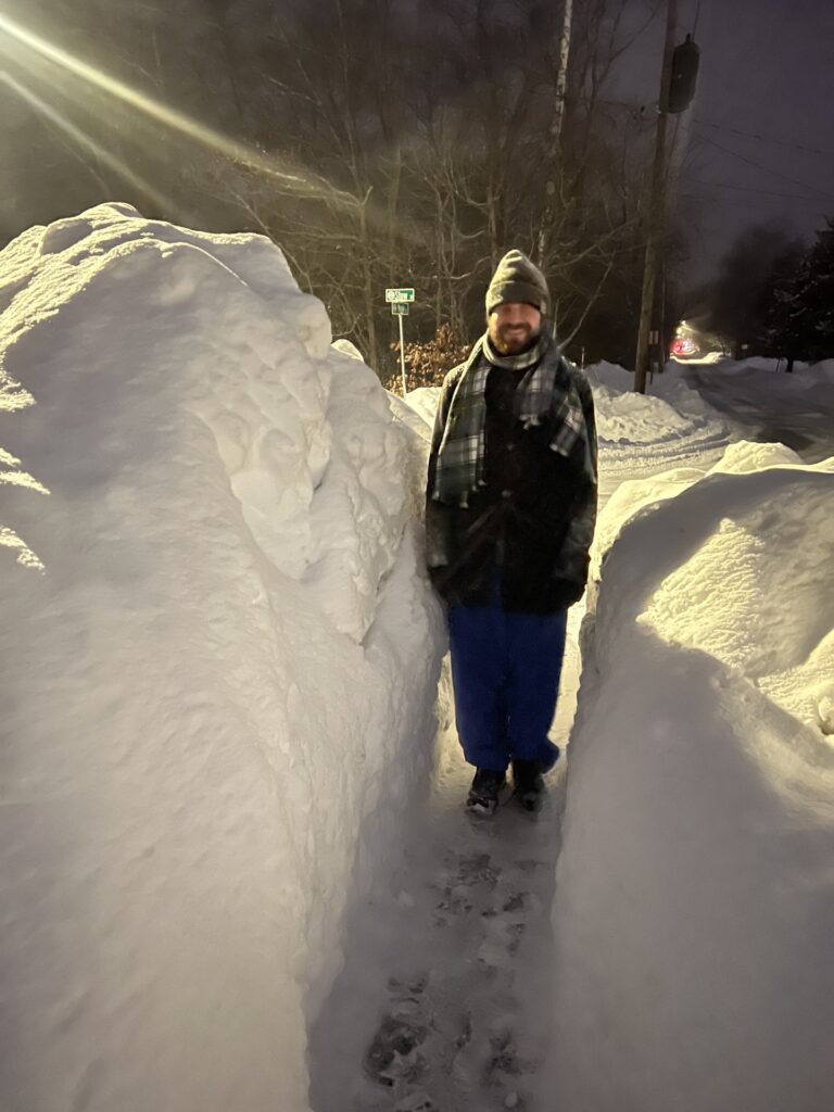 A man stands in a shoveled path. On one side, the snow is a little taller than his hat.